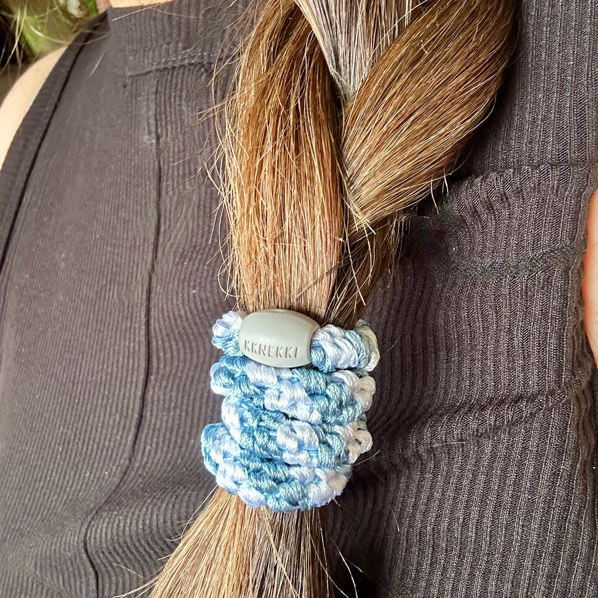 A close-up of a person's braided brown hair secured with a KKNEKKI Sea-Blue and White striped hair tie featuring a branded 'KKNEKKI' bead.