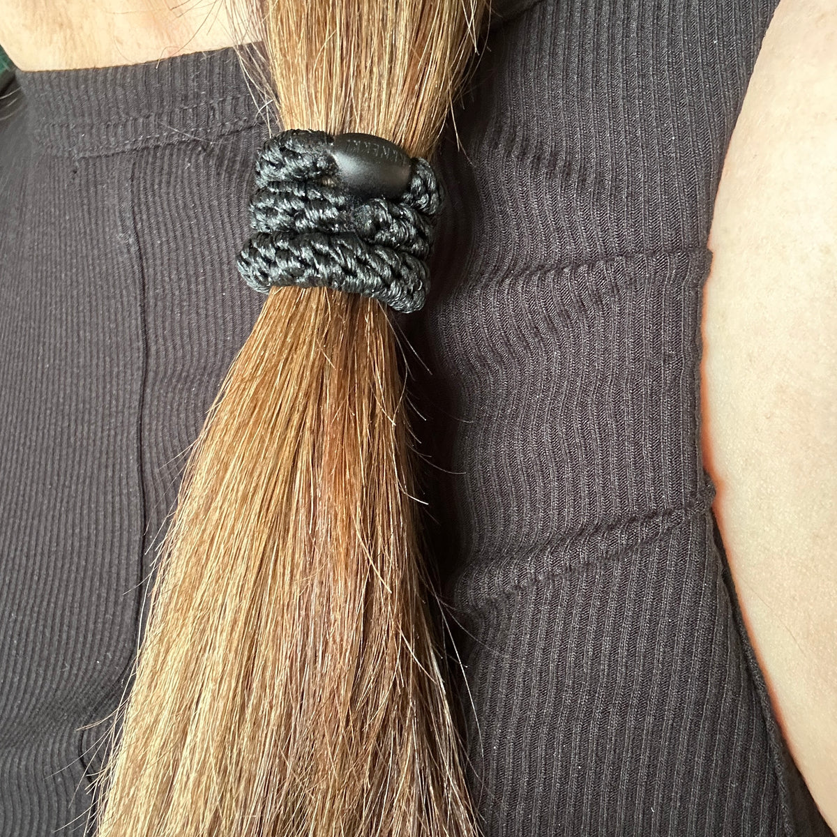 A close-up of a person's hair tied back in a low ponytail with a KKNEKKI Black Glitter Hair tie featuring a branded 'KKNEKKI' bead, worn over a dark ribbed top.