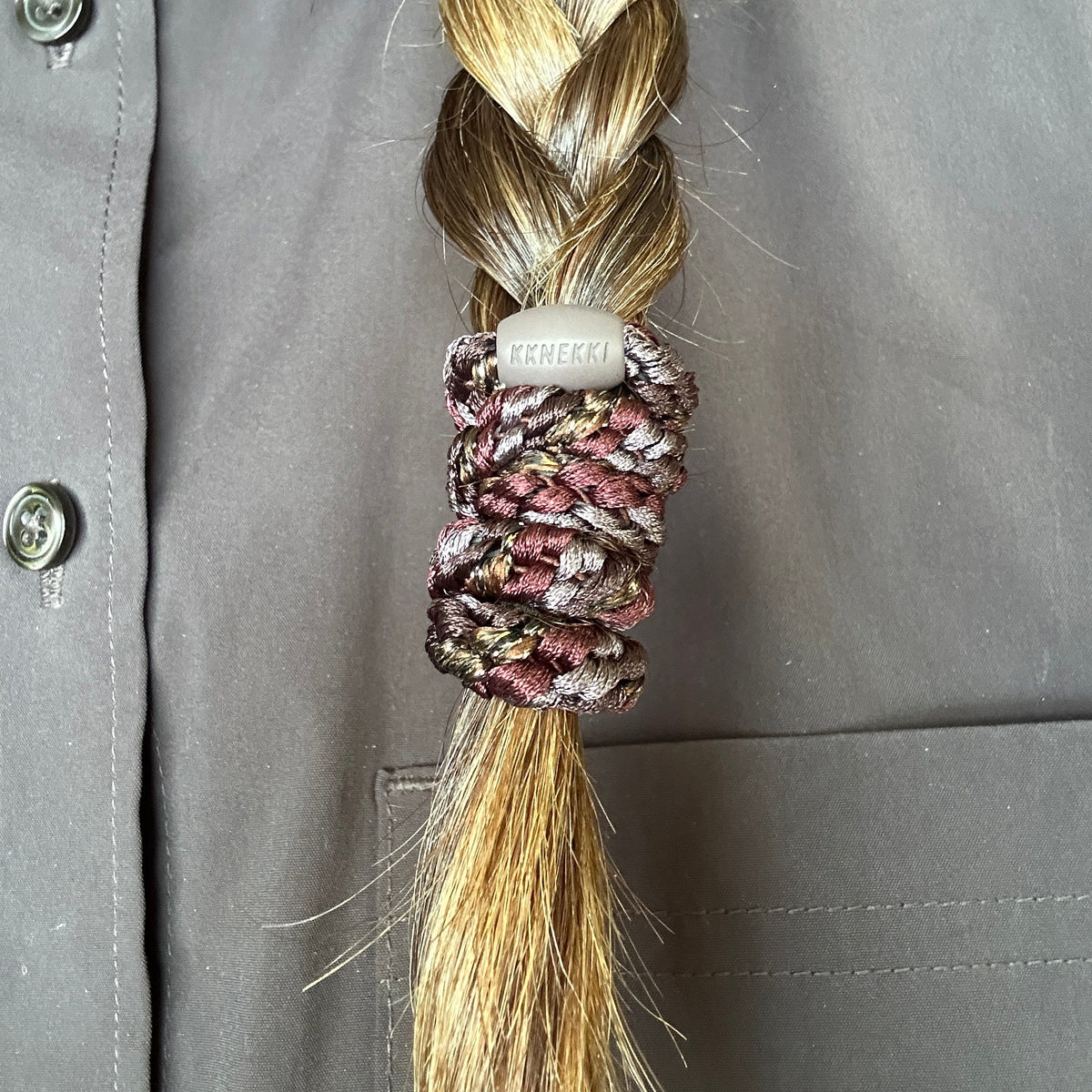 A close-up of brown braided hair secured by a KKNEKKI hair elastic in the color Mix Cedar Coffee Glitter, a striped luxury hair tie with subtle glitter strands, against a brown button-up shirt.