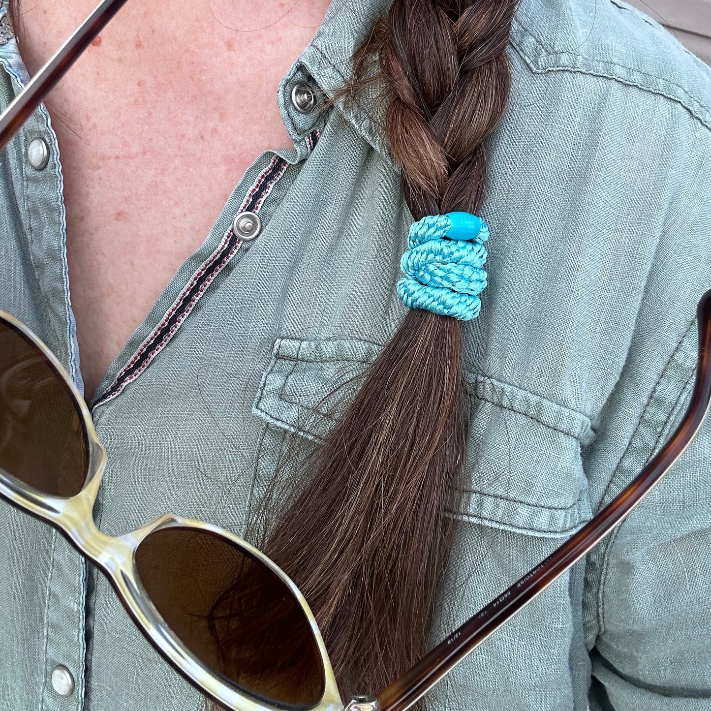 KKNEKKI Lake Blue hair elastic worn in a brown braid and over a green linen shirt with snap closures and near sunglasses