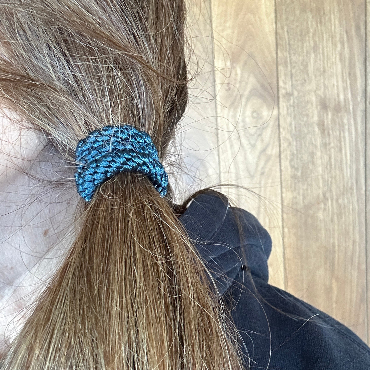 A close-up lifestyle shot of a person's brown hair tied back in a low ponytail with a luxury woven KKNEKKI Petrol blue colored hair elastic set against a wooden paneled wall.