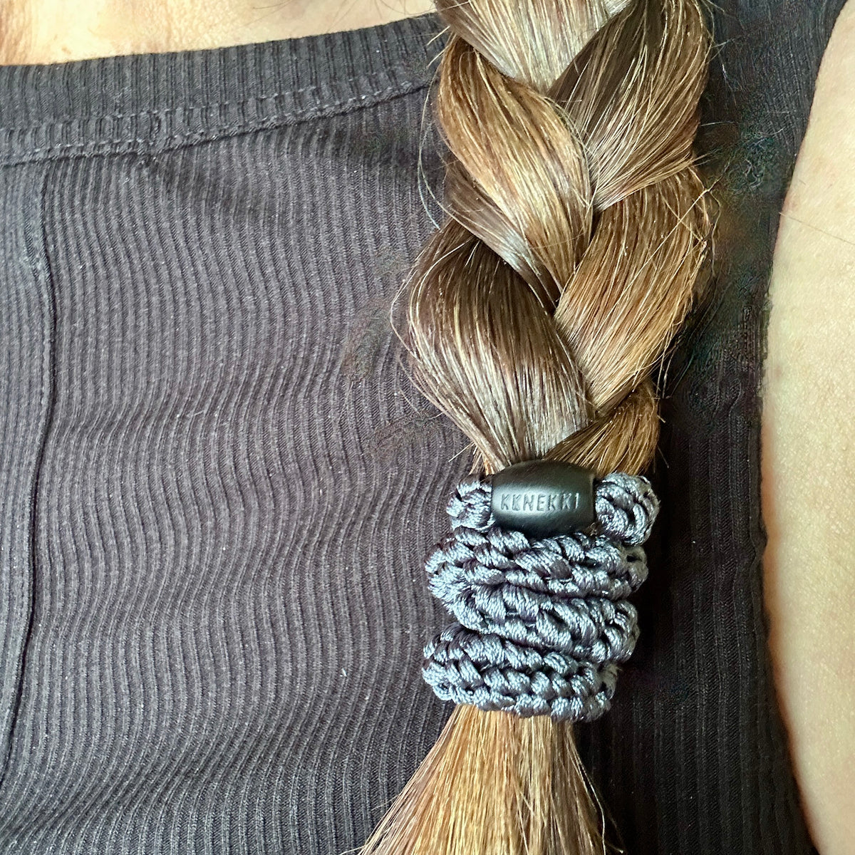 A close-up of a person's braided brown hair secured by a KKNEKKI hair elastic with dark grey branded 'KKNEKKI' bead visible. The person is wearing a dark ribbed tank top.