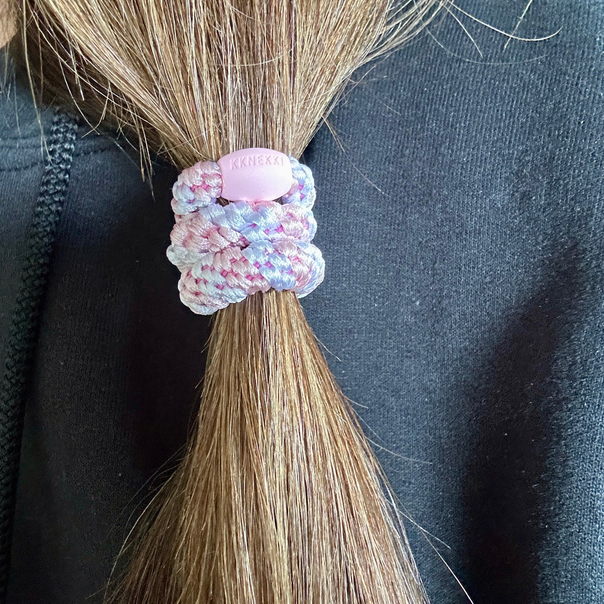 A close-up of a person's light brown hair tied in a low ponytail with a KKNEKKI hair tie in the color Light Pink & White, worn over a black top.
