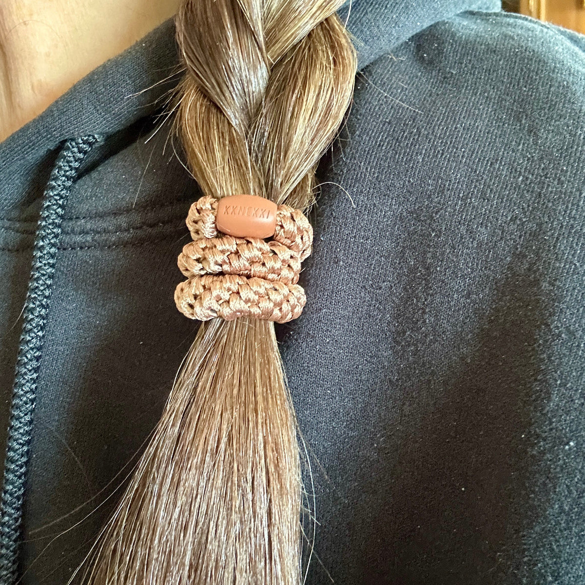 A close-up of a person's light brown hair tied in a low ponytail with a KKNEKKI hair elastic in the color Nougat, worn over a black hoodie.