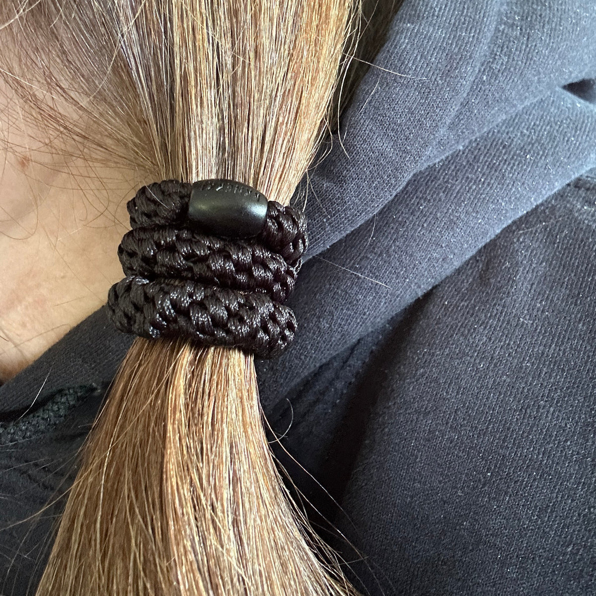A close-up of a person's light brown hair tied in a side ponytail with a KKNEKKI hair elastic in the color Black, worn over a black hoodie, branded bead visible.