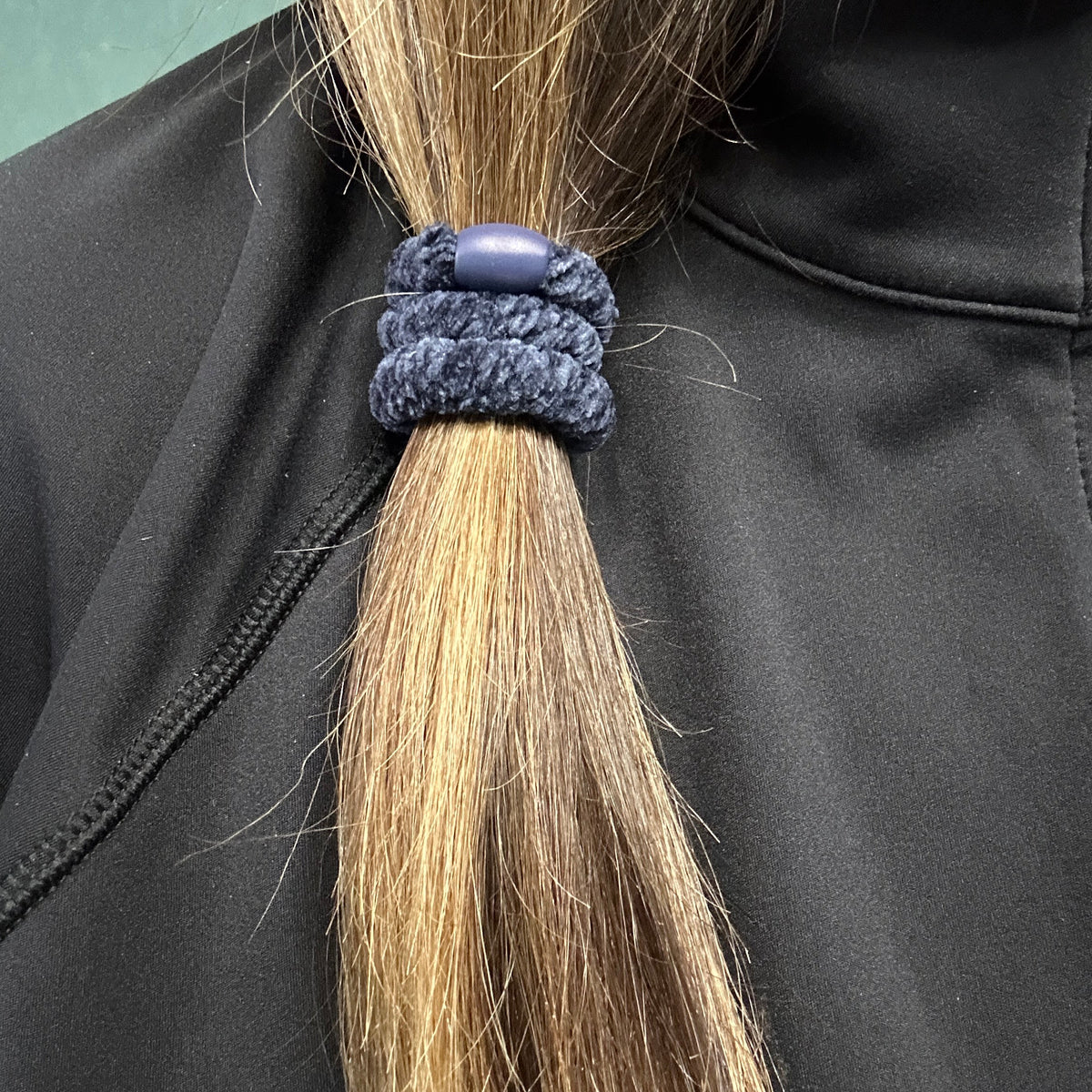A close-up of a person's light brown hair tied in a side ponytail  with a KKNEKKI hair tie in the color Navy Velvet, worn over a black jacket.