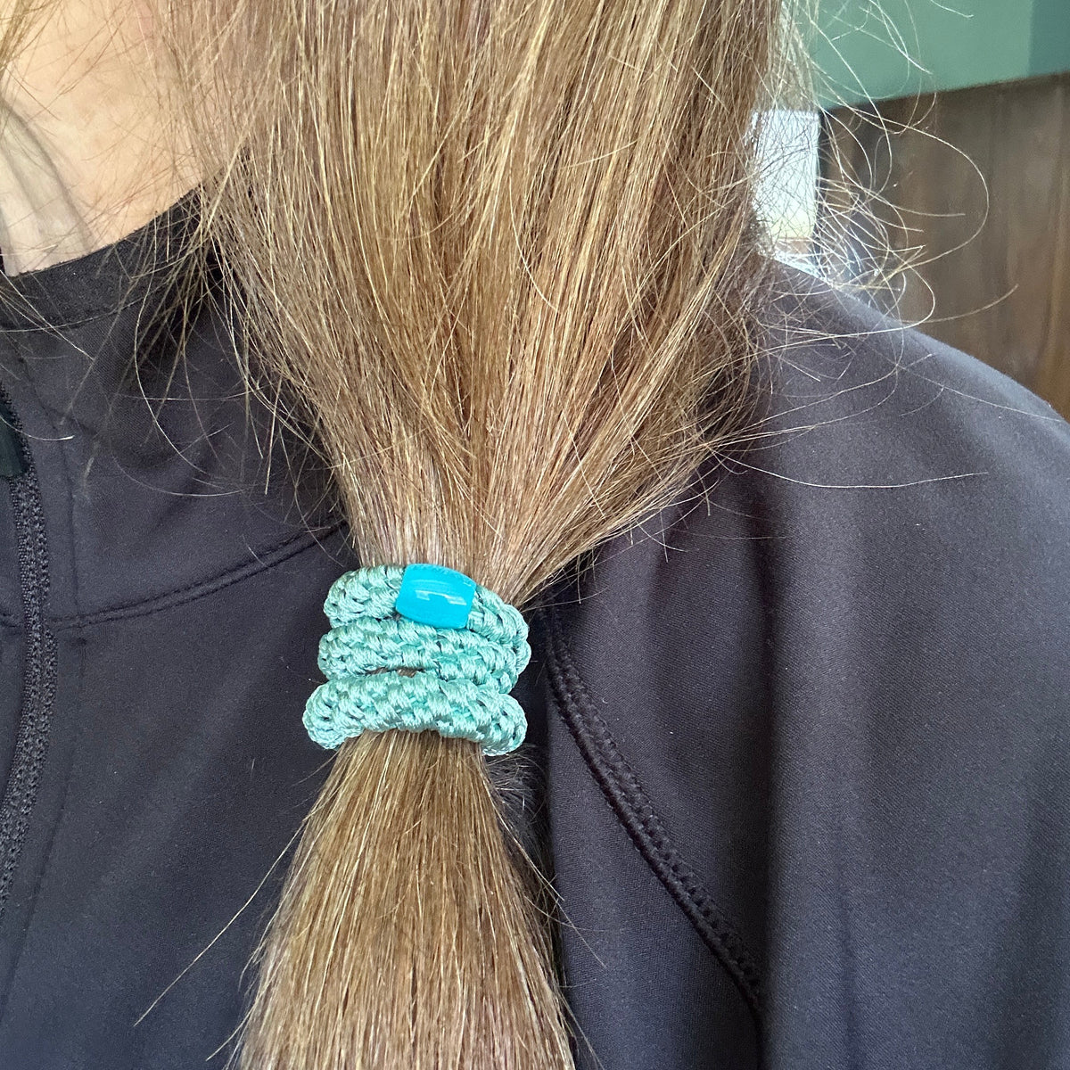 A close-up of a person's long light brown hair tied back in a low ponytail with an authentic original KKNEKKI  woven hair elastic in Teal..