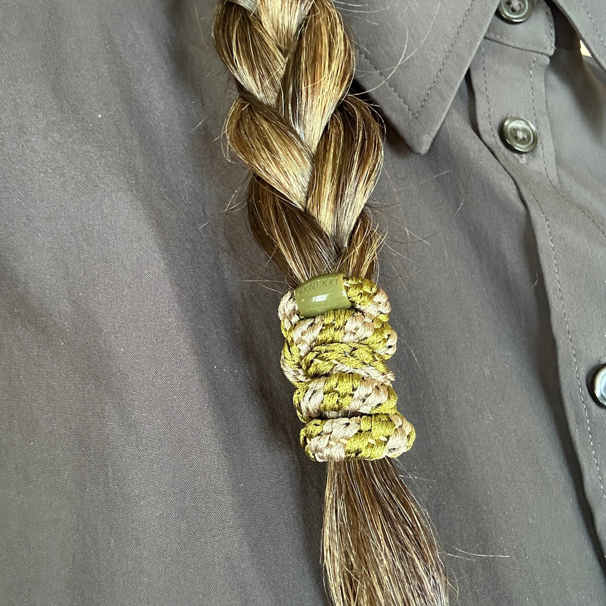 A close-up of braided hair secured by a luxury olive green and beige KKNEKKI hair elastic in the color Camo, against a brown button-up shirt.