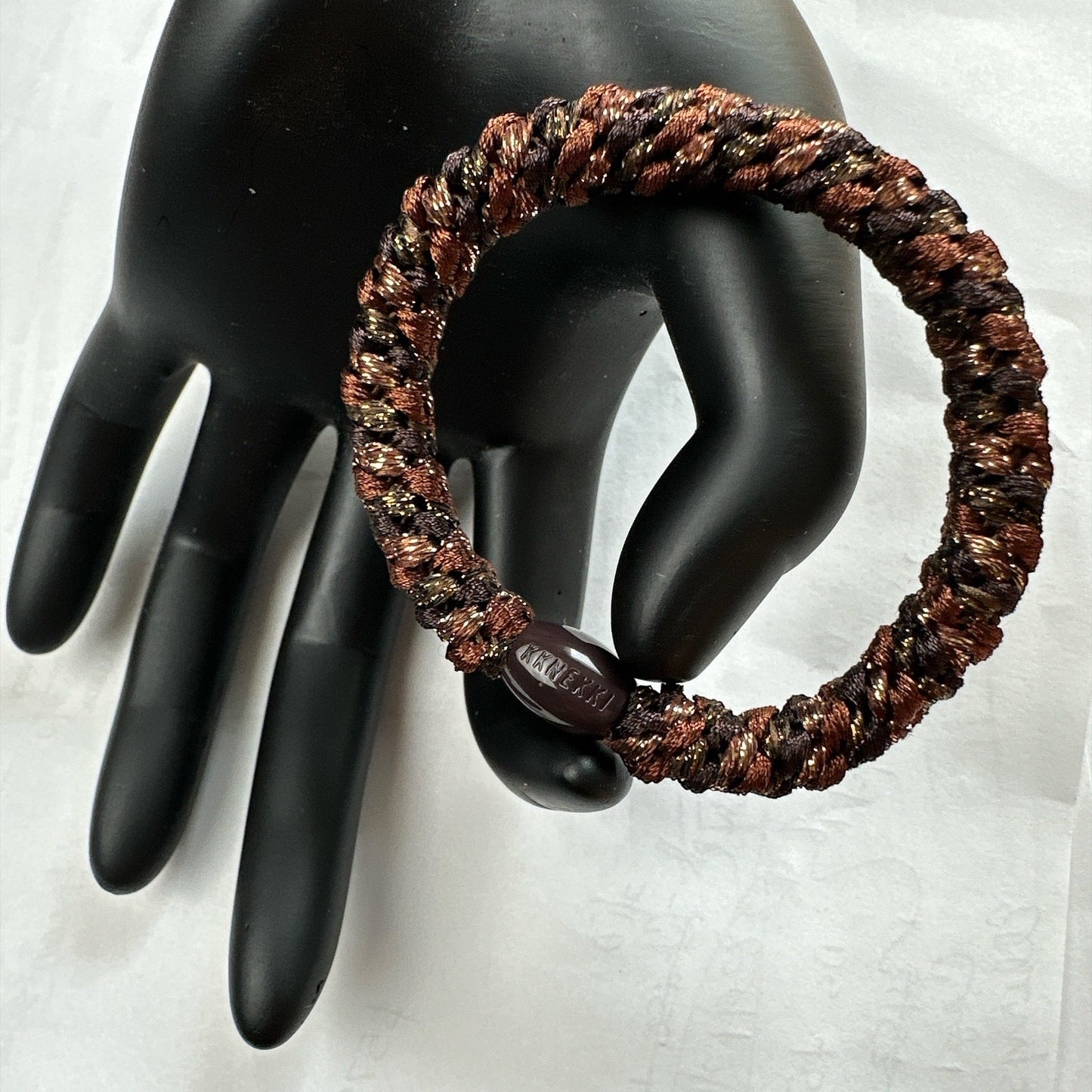 KKNEKKI Brown Stormy hair tie braided elastic with golden threads on a black mannequin hand against a white background
