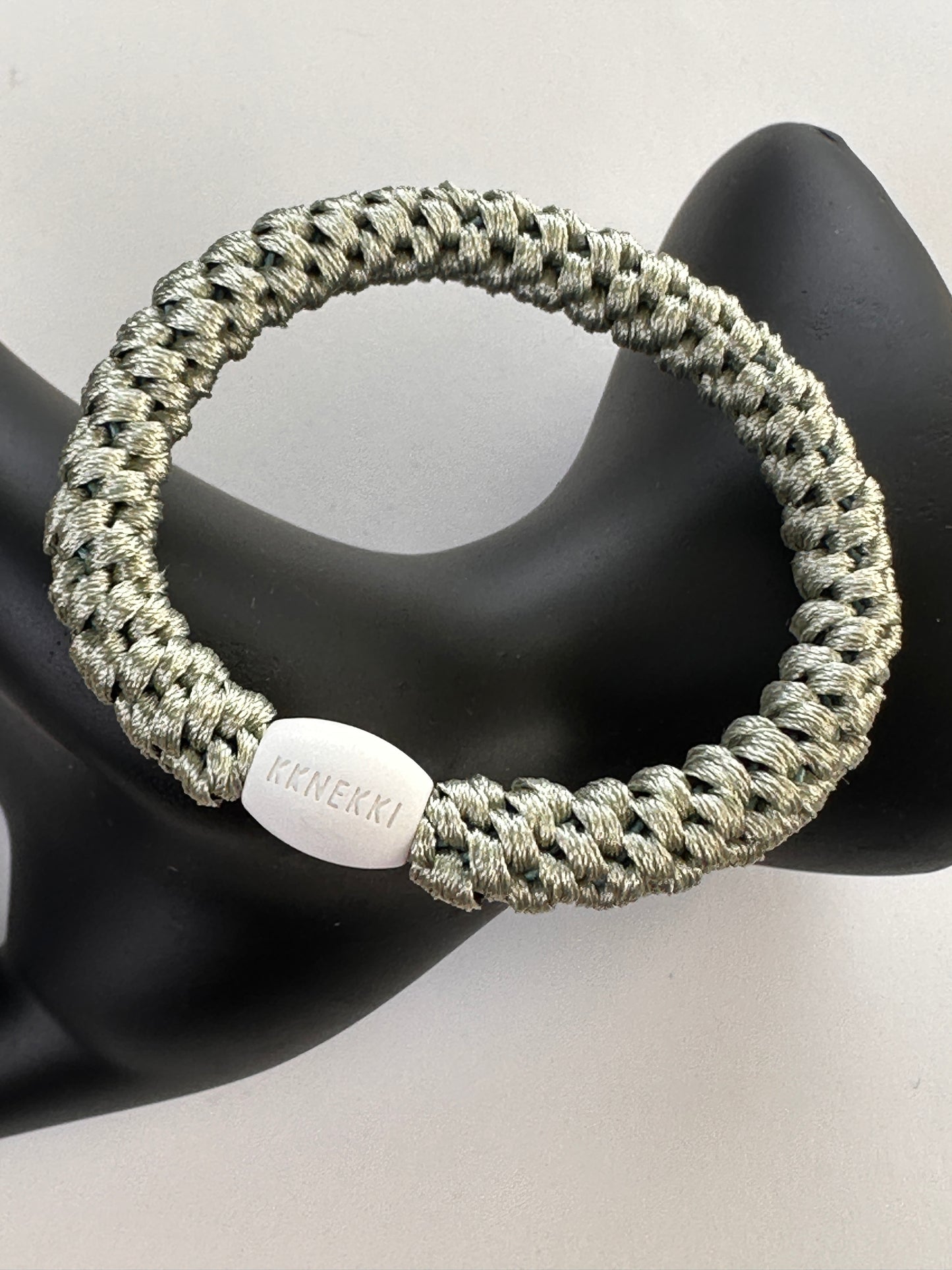 KKNEKKI Dusty Lind Green hair elasticon a black model hand with a white background showing branded bead