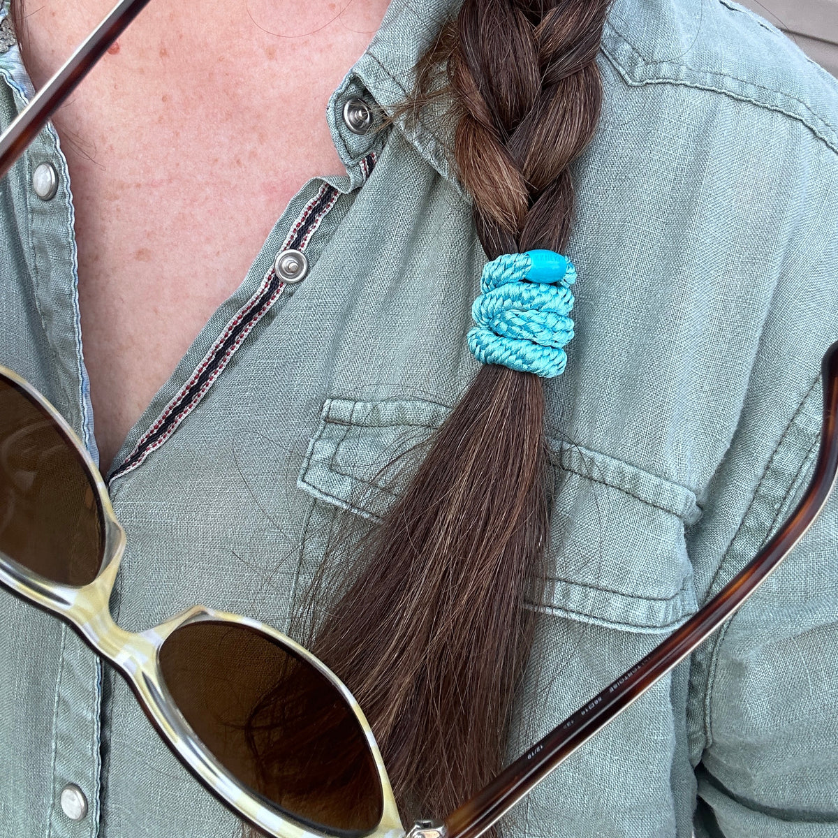 KKNEKKI Lake Blue hair elastic worn in a brown braid and over a green linen shirt with snap closures and near sunglasses