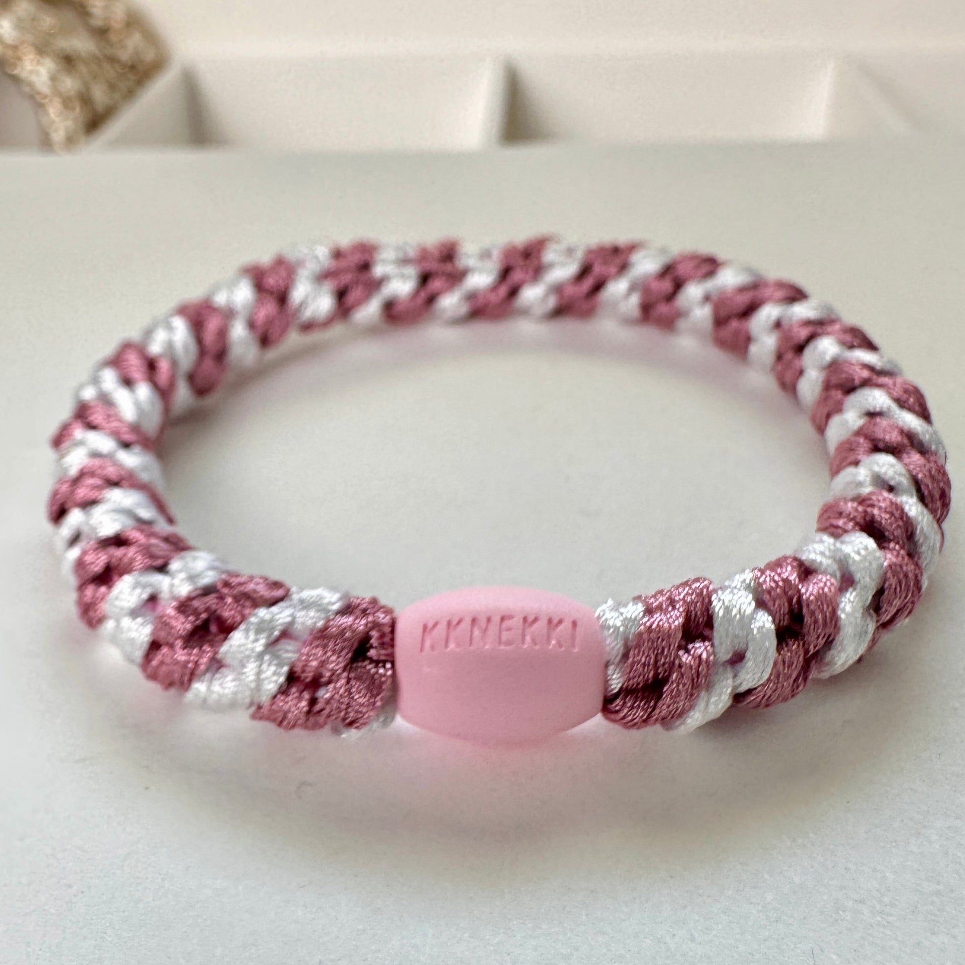 KKNEKKI Old Pink & White Stripe hair tie laying slightly sideways on a white box  and you can see the branded bead