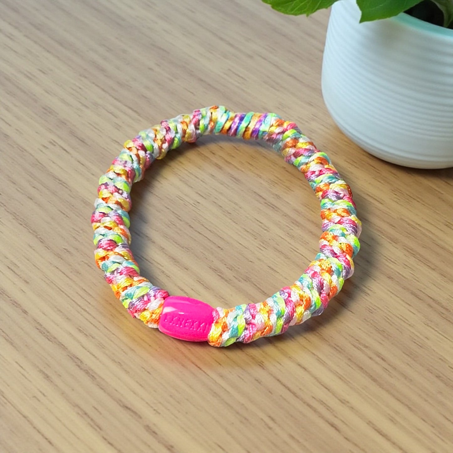 KKNEKKI Rainbow hair tie on wood desk top near plant in white container. Branded bead clearly in view.