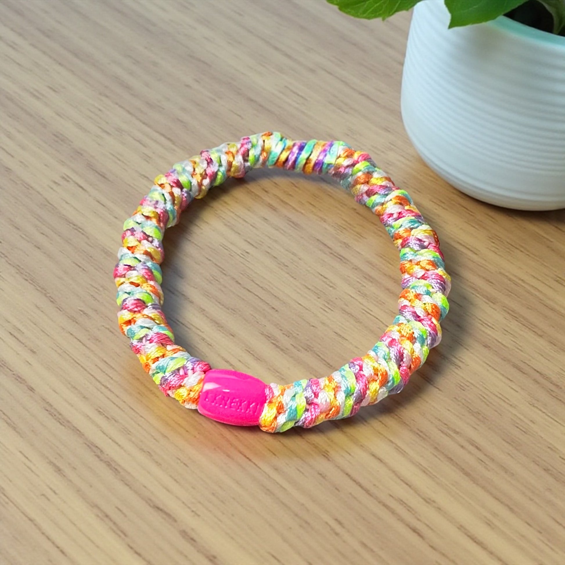KKNEKKI Rainbow hair tie on wood desk top near plant in white container. Branded bead clearly in view.