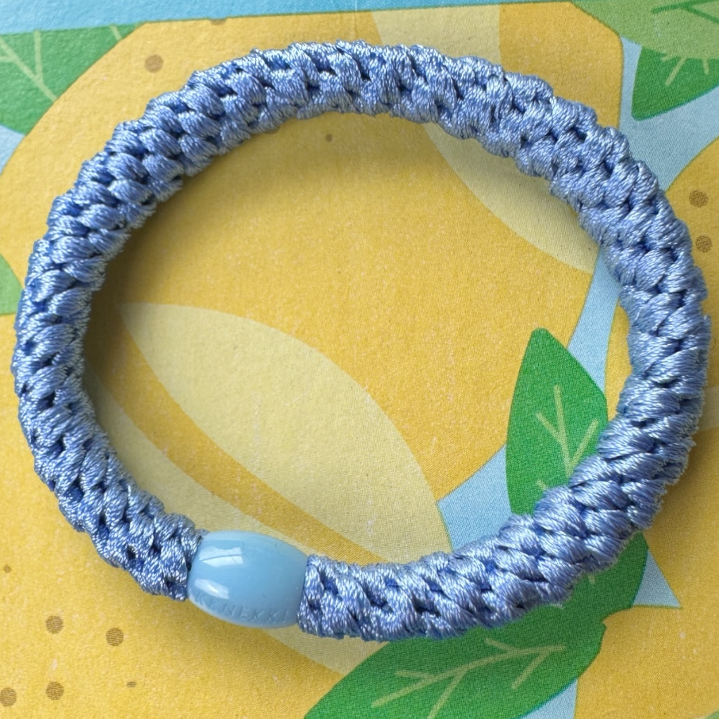 KKNEKKI Robin's Blue hair elastic sitting on a colorful yellow, green, and blue background.