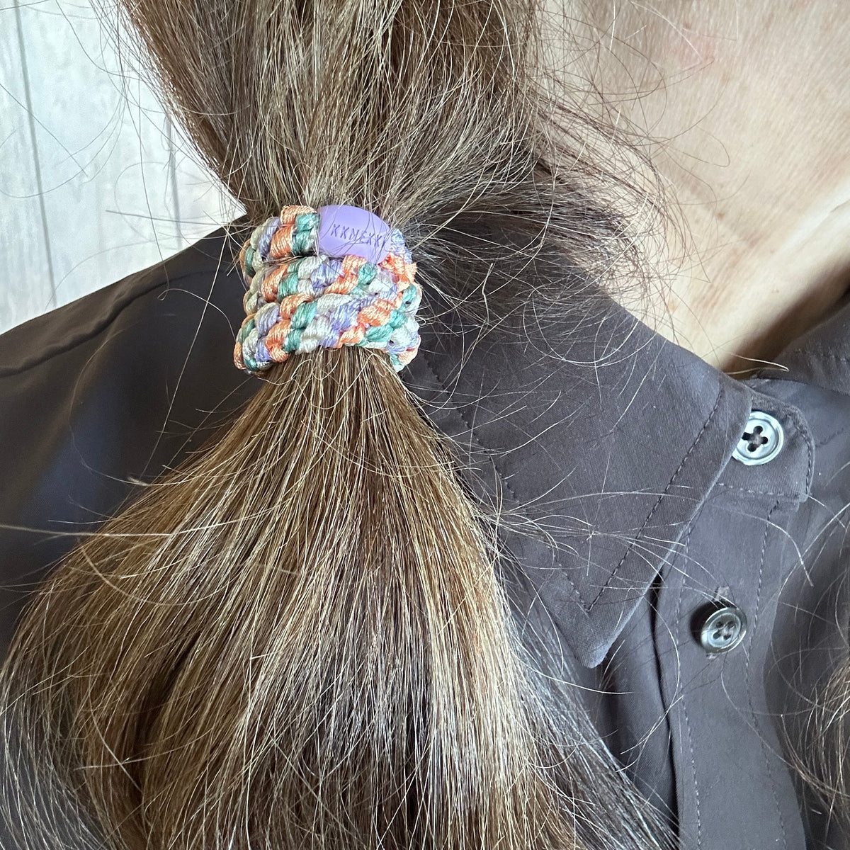 KKNEKKI woven with 60+ strands luxury hair elastic in the color Mix Mermaid holding a person's brown-haired side ponytail, branded bead visible, against a dark brown shirt.