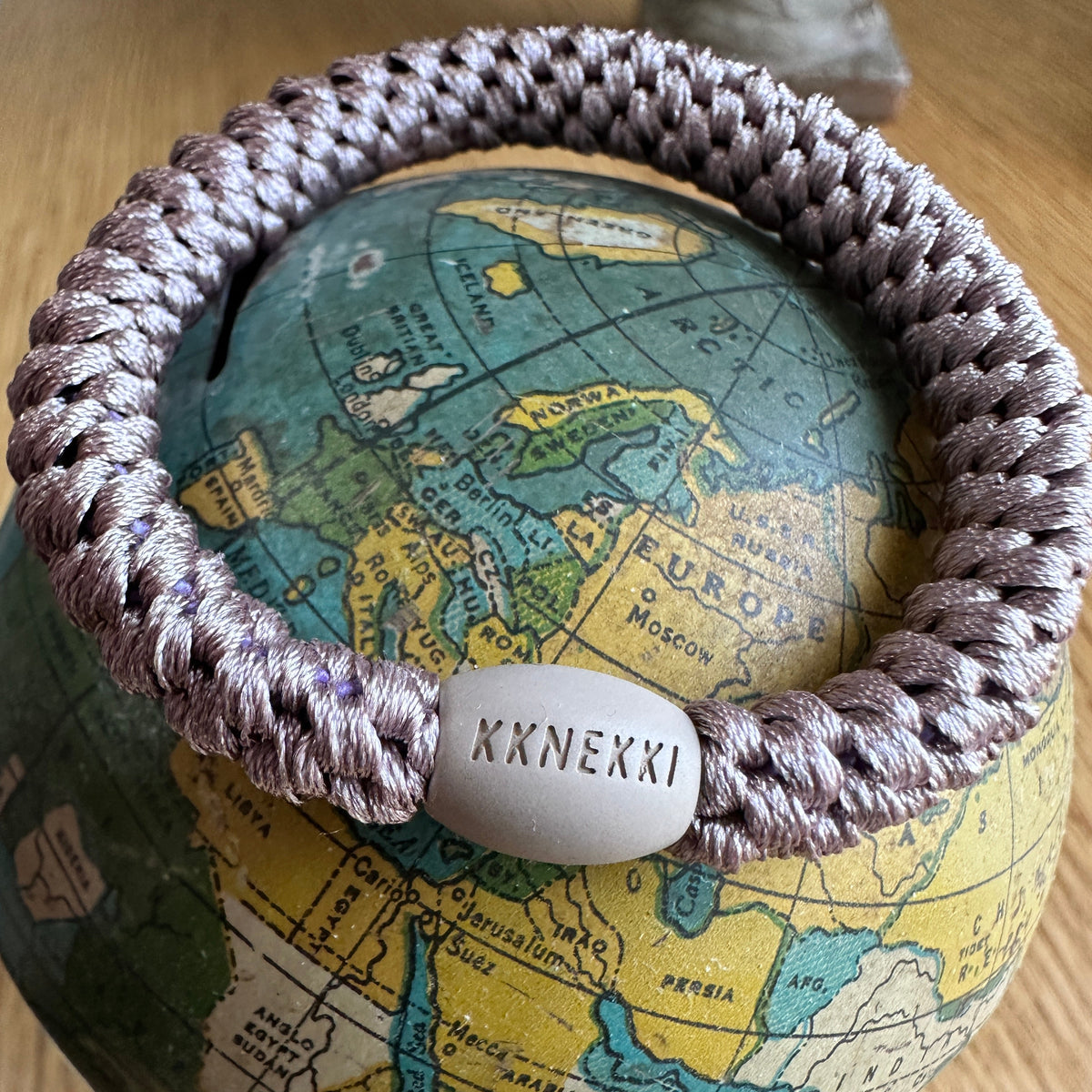 Lifestyle photo of a closeup of and authentic original KKNEKKI hair tie in the color Dove perched on top of a green, yellow, and blue antique globe bank sitting on a woodgrain desk, branded bead clearly visible.