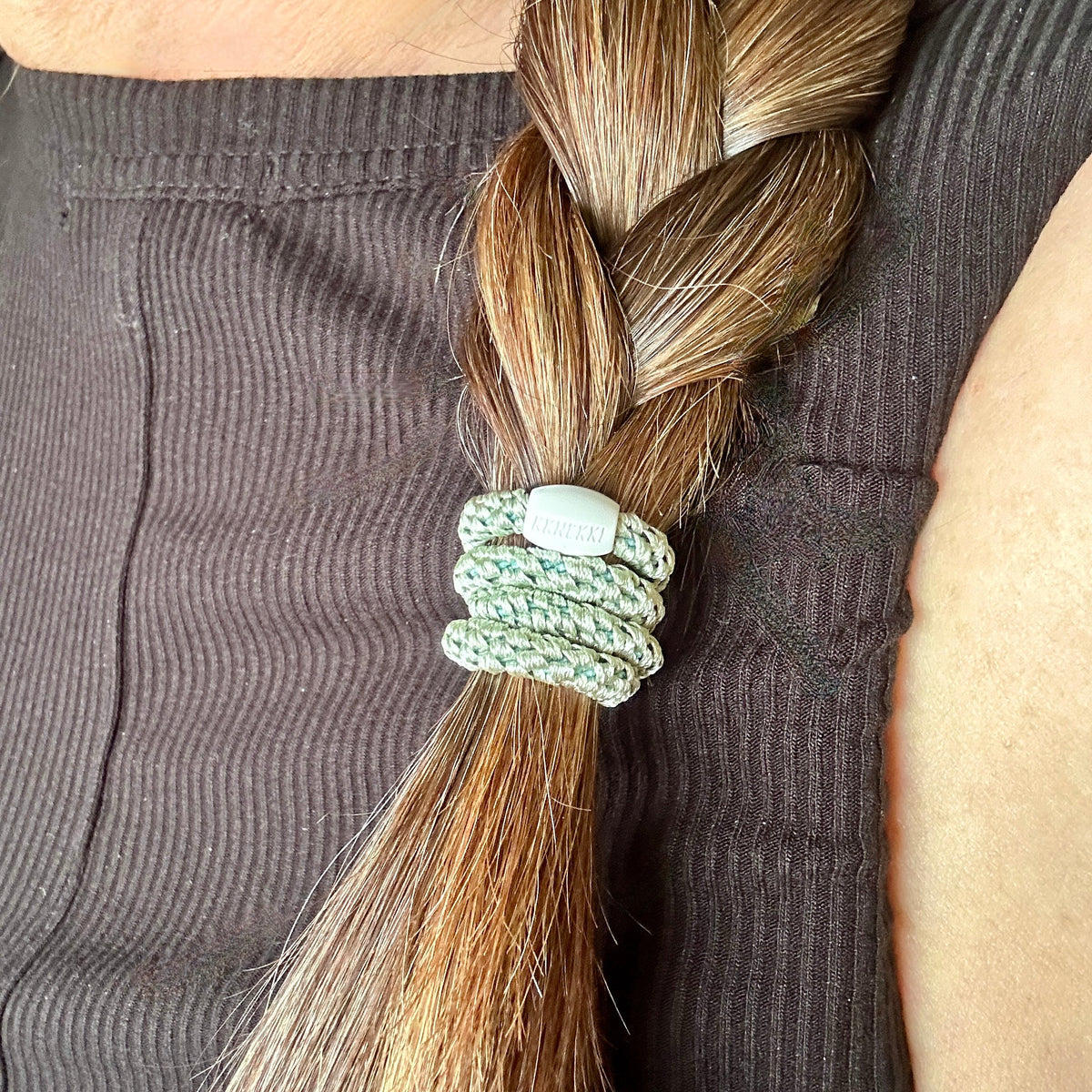 Photograph of a person with long brown hair secured by a KKNEKKI Slim Dusty Lind colored hair elastic against their dark ribbed shirt, 'KKNEKKI' branded bead visible.